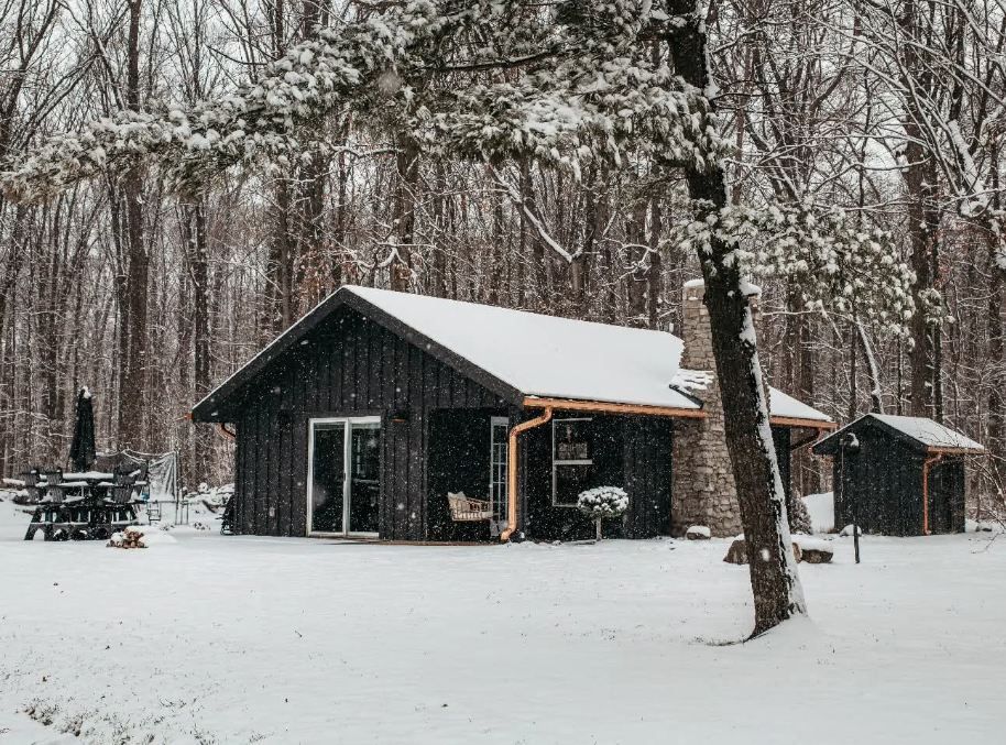 Snow-covered cabin in a wooded forest with trees and a small shed nearby