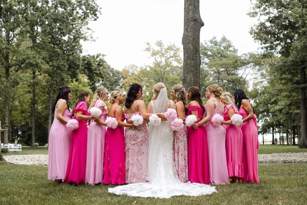 Bridal party in pink dresses standing with bride in white outdoors near trees