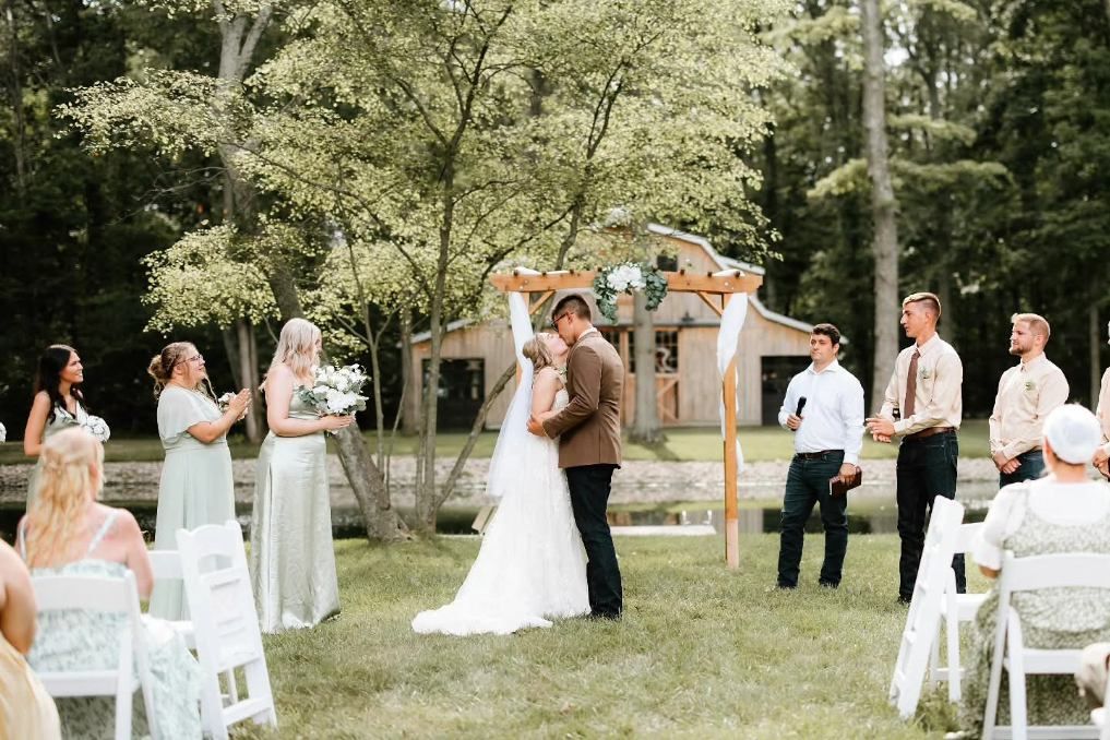 Outdoor wedding ceremony with bride and groom under a wooden arch, guests seated on a lawn amid trees