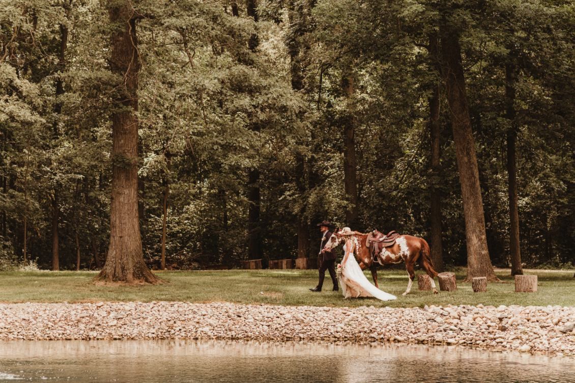 Bride and groom holding hands by a lake in a wooded park, with a white wedding dress train trailing behind.