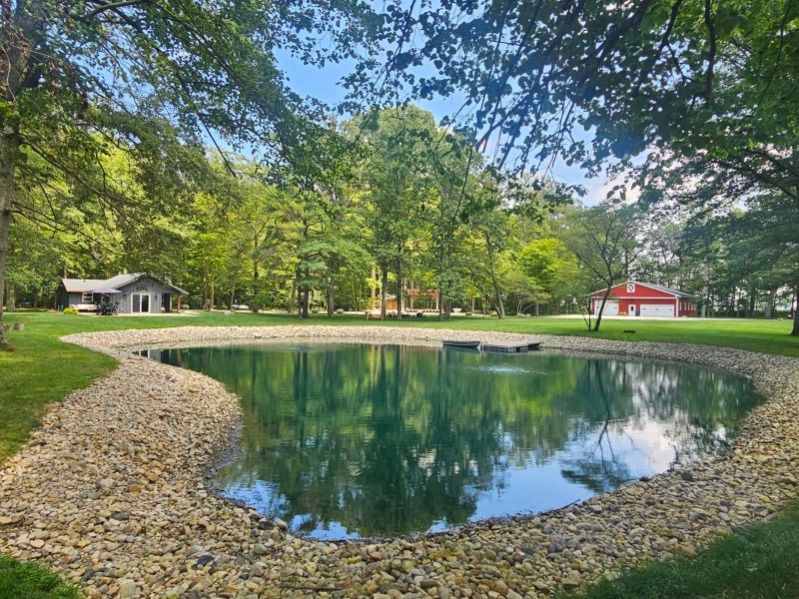 Wooded park with a small pond reflecting trees and two small buildings in the background.