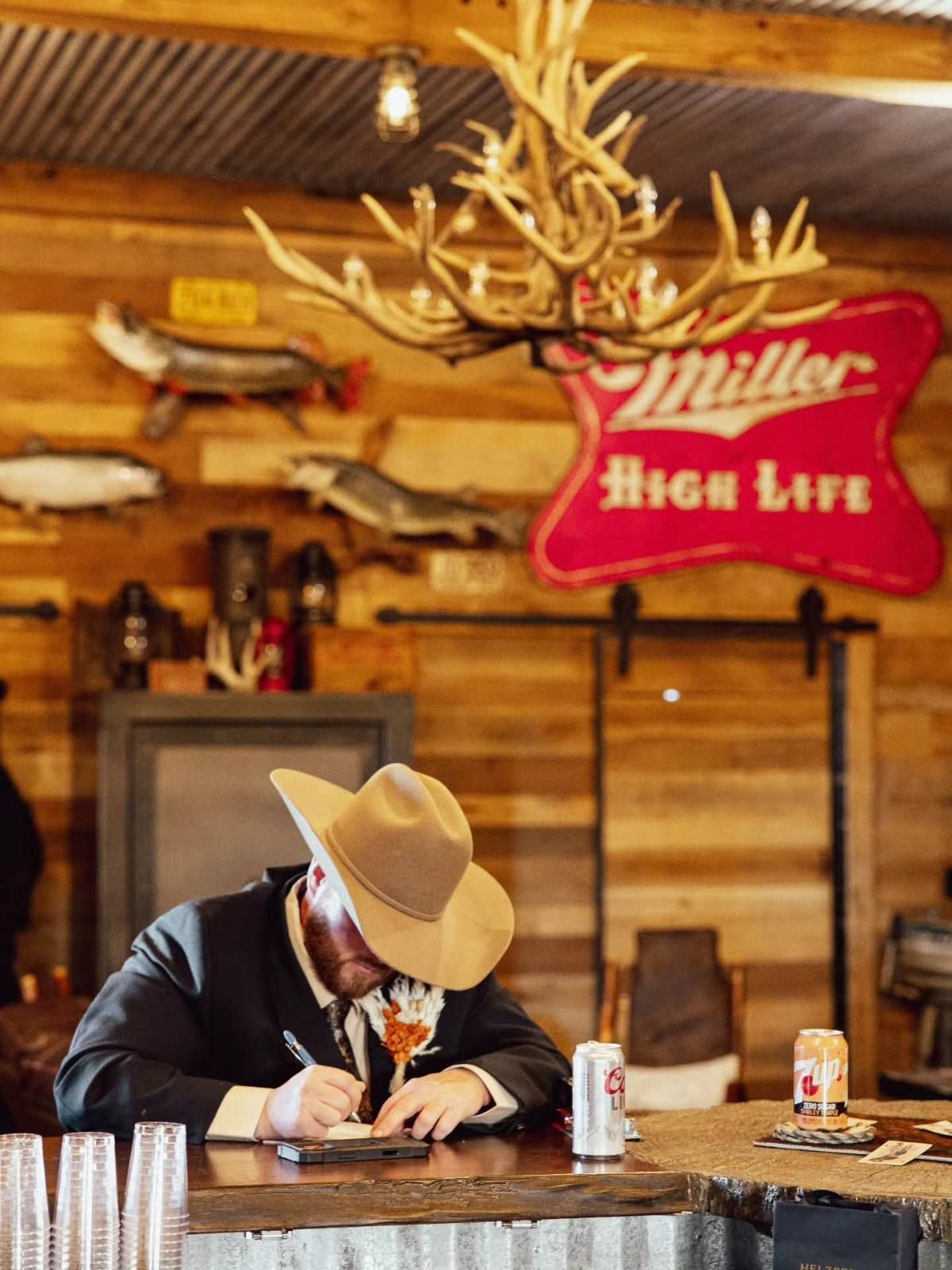 Cowboy-hatted man writing at a bar under a Miller High Life sign