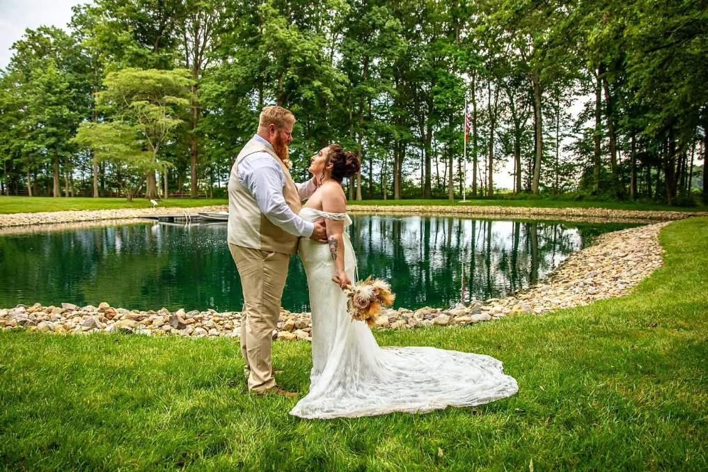 Bride and groom stand by a pond in a green park, embracing on their wedding day.