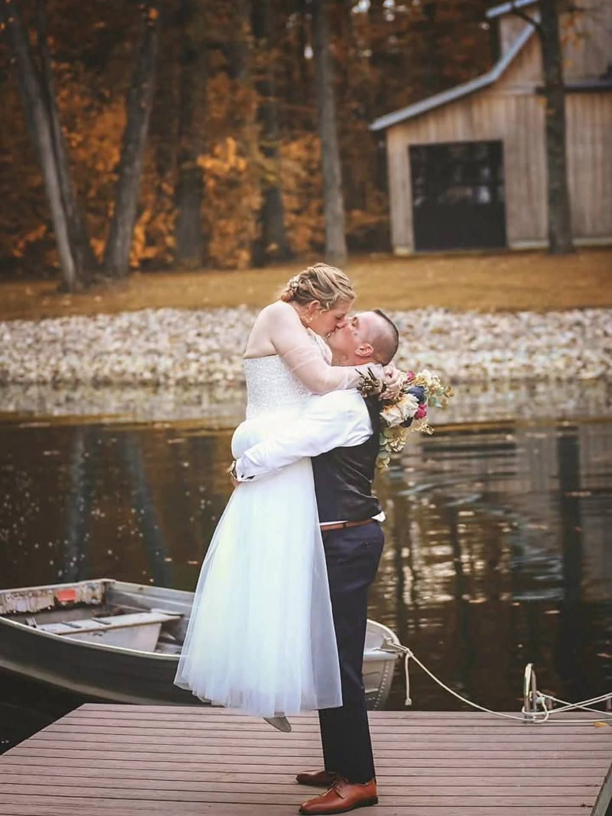 Bride and groom kiss on a dock by the water, with a small boat and autumn trees in the background.