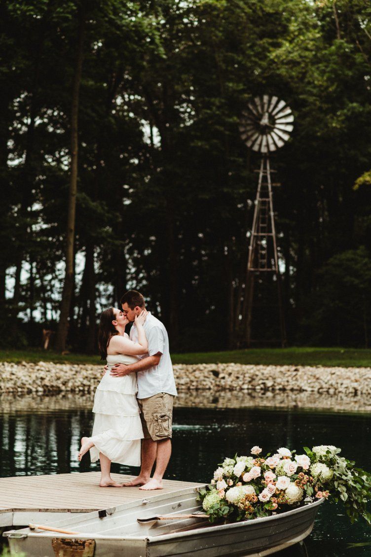 Couple embracing on a wooden dock by a pond, with a windmill and trees in the background.