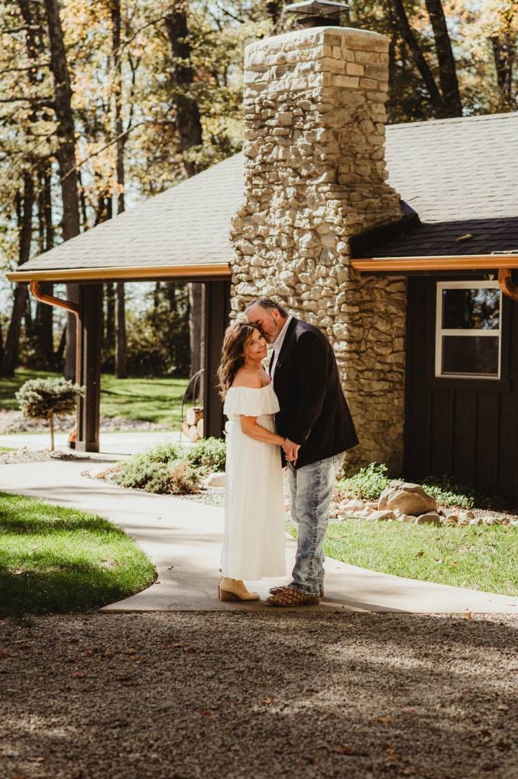 Couple in wedding attire kissing beside a stone chimney at a rustic cabin in the woods