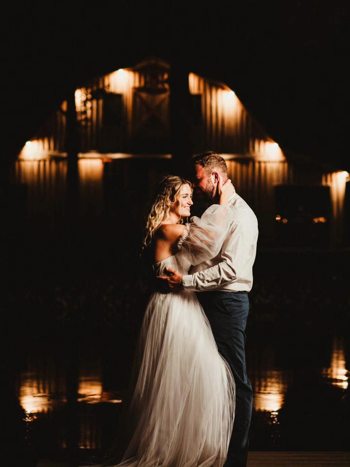 Couple embracing in formal attire on a dimly lit stage with warm lights reflected behind them