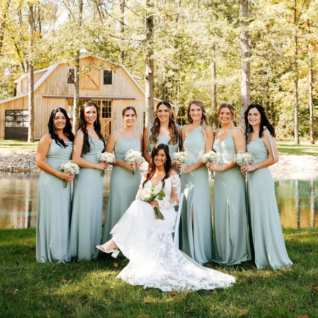 Bridesmaids in sage dresses pose with bride in white gown in front of a rustic barn by a pond.