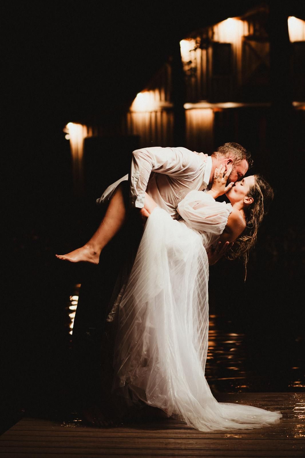 Couple kissing in a romantic dip pose by a dark, candlelit staircase, bride in a white gown.