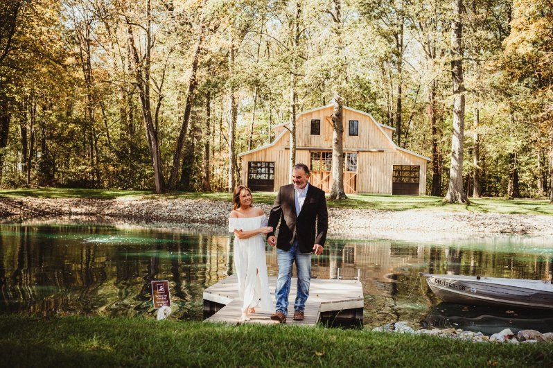 Couple standing on a dock by a pond, with a small house and wooded trees in the background.