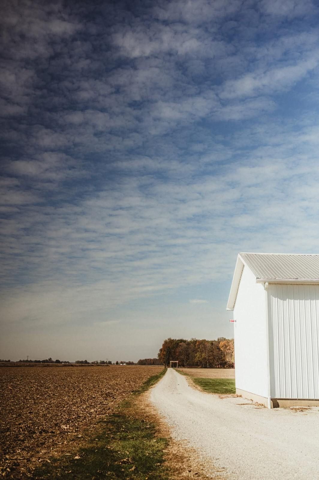 Rural dirt road beside a white barn under a cloudy blue sky