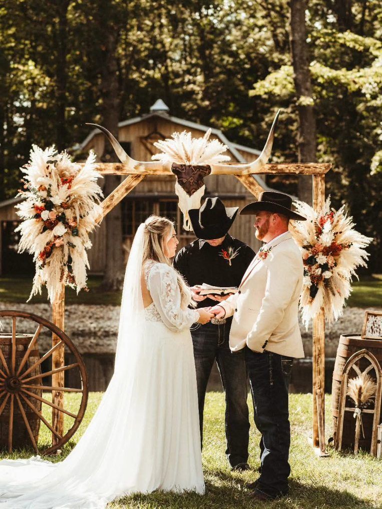 Wedding couple and officiant under a floral arch in a wooded outdoor ceremony