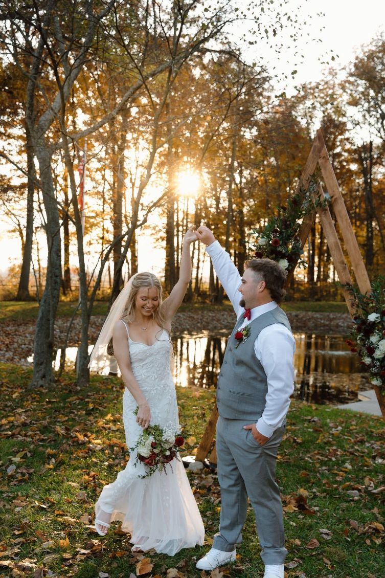 Newlyweds holding hands in a sunlit autumn park, raising joined arms beside a pond