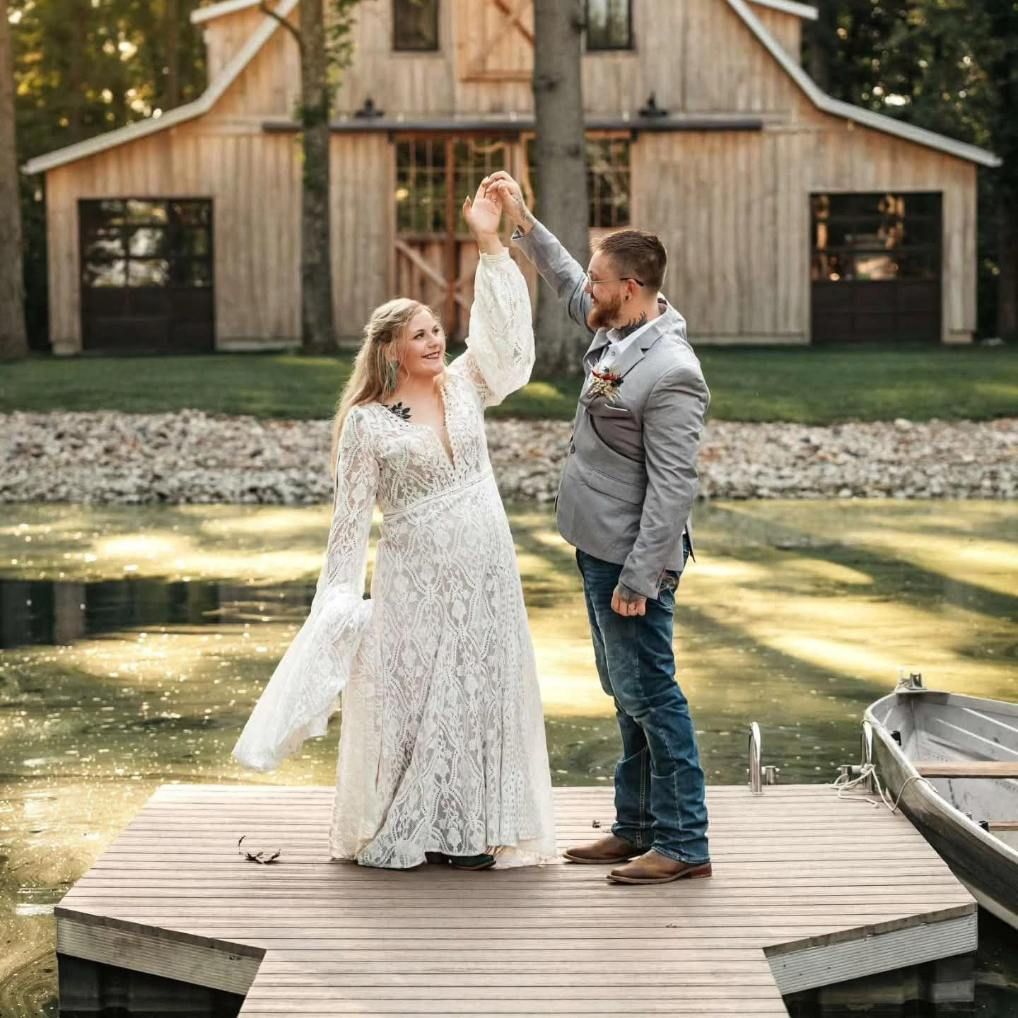 Couple dancing on a wooden dock by a lake, with a rustic barn in the background.