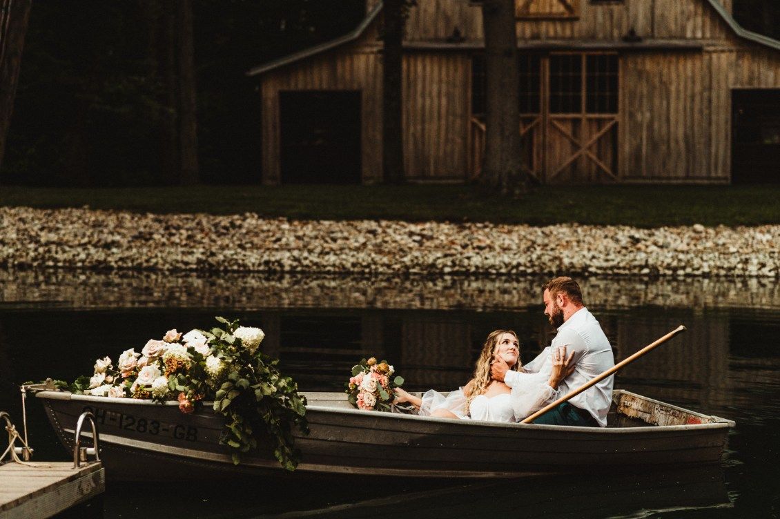 Bride and groom in a rowboat beside a rustic boathouse, with white flowers and a calm canal