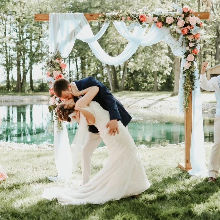 Couple sharing a kiss under a floral arch by a lakeside at an outdoor wedding ceremony