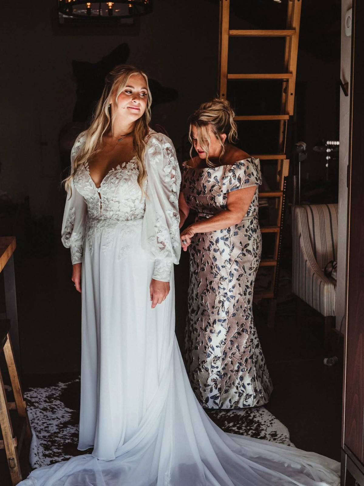 Woman in a white dress and veil stands beside another in a floral gown in a dim indoor room.
