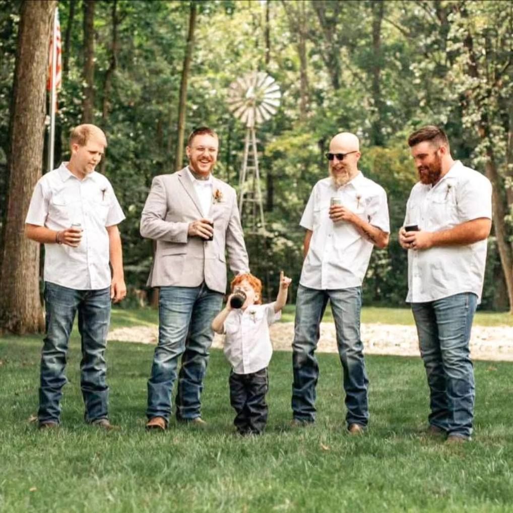 Five people stand on grass in a wooded park, one child lifted between four adults, all holding drinks.