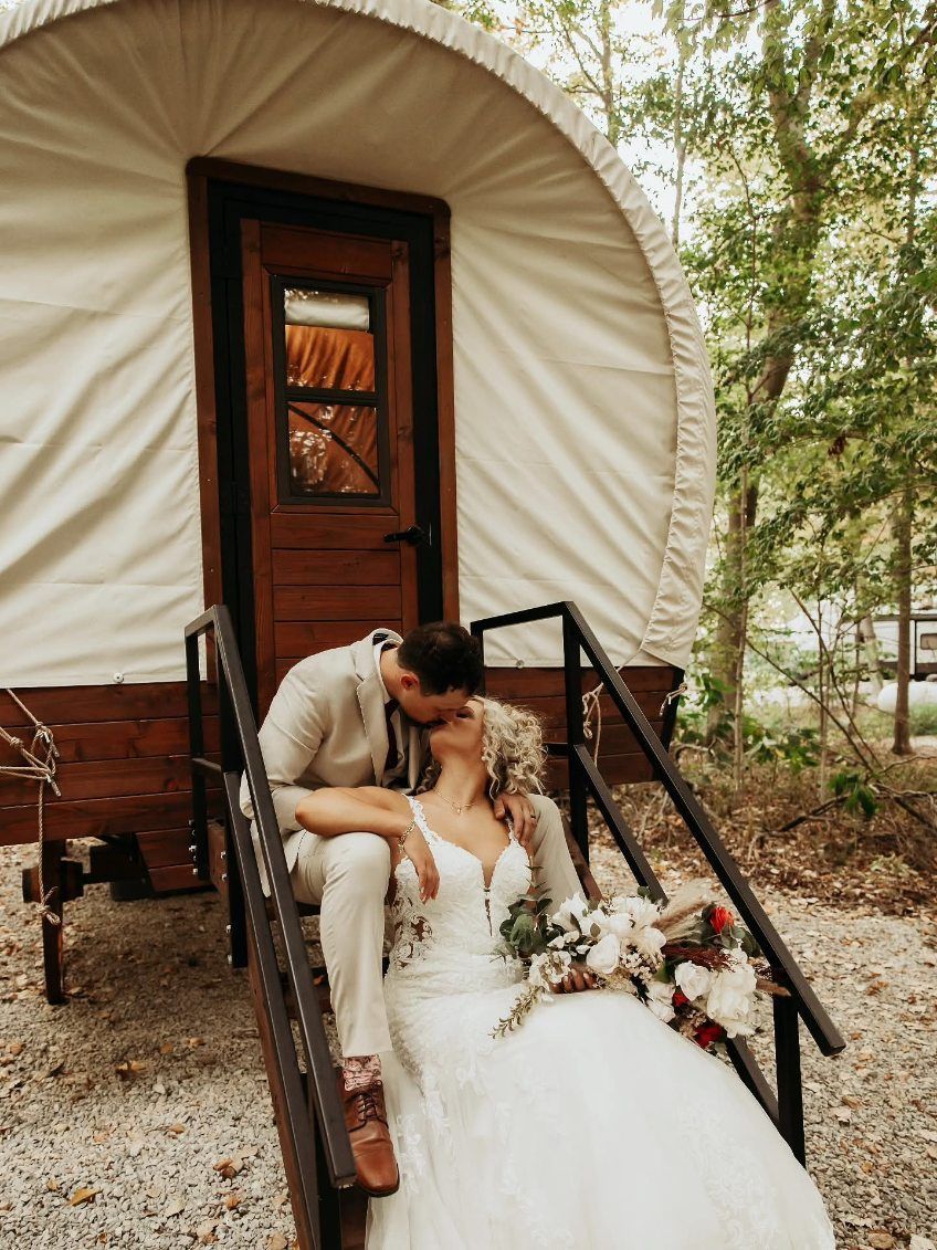 Bride and groom sitting on wagon steps outside a white covered wagon, sharing a kiss with flowers nearby