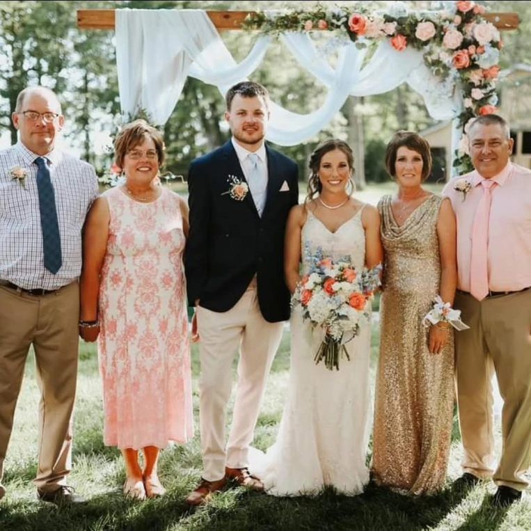 Wedding party standing under floral arch outdoors, with bride and groom centered among family members