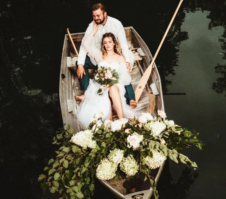 Bride and groom in a rowboat on dark water, surrounded by white flowers
