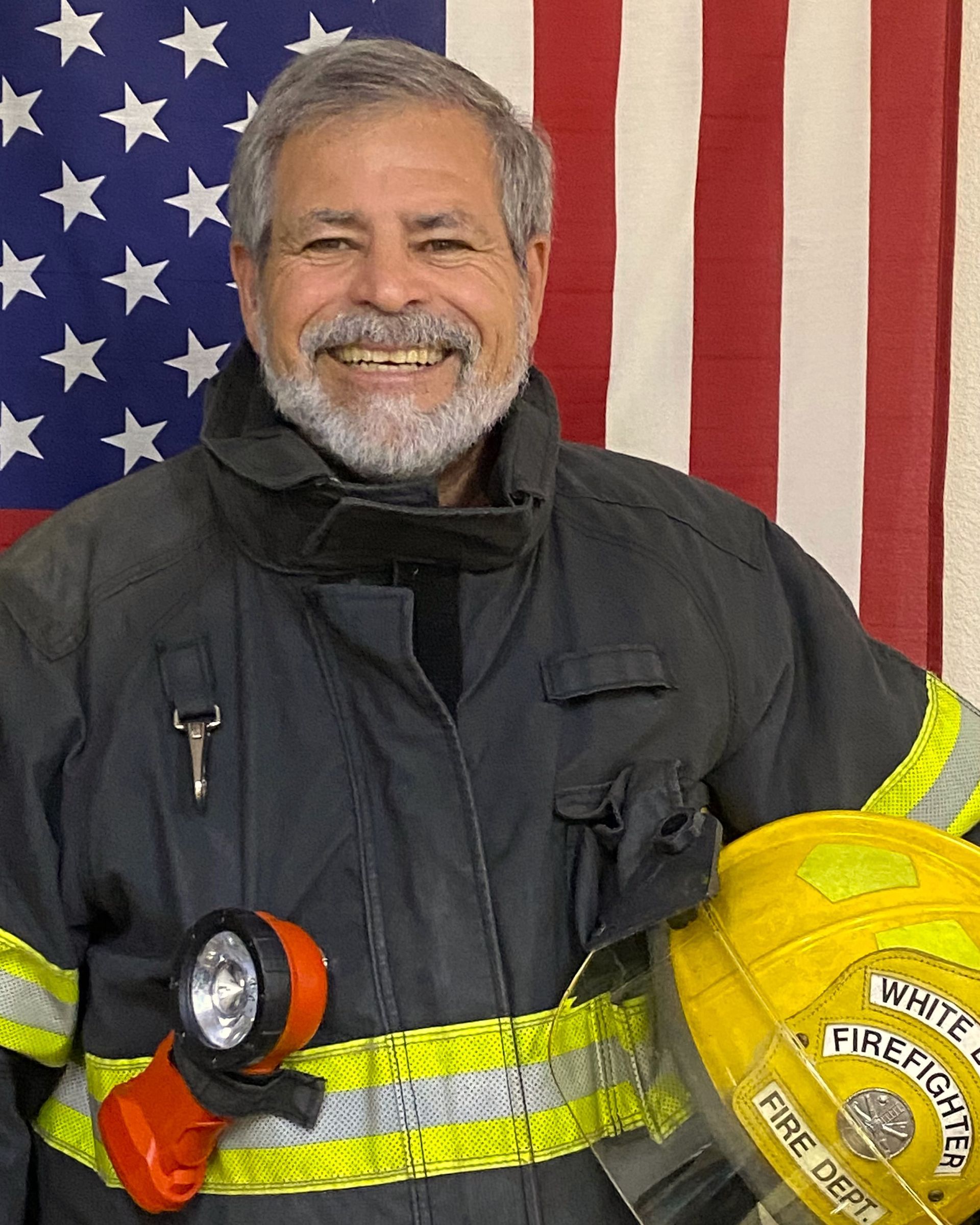A man in a fireman 's uniform is holding a helmet and a flashlight in front of an american flag.