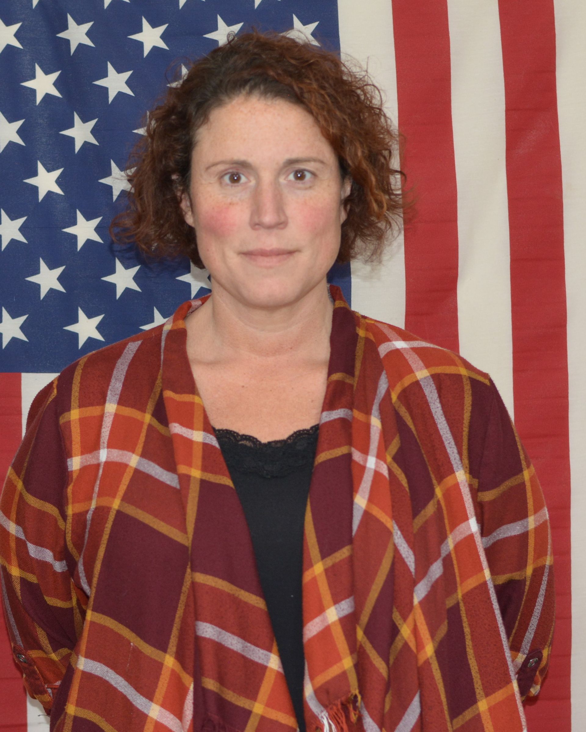 A woman in a plaid jacket stands in front of an american flag
