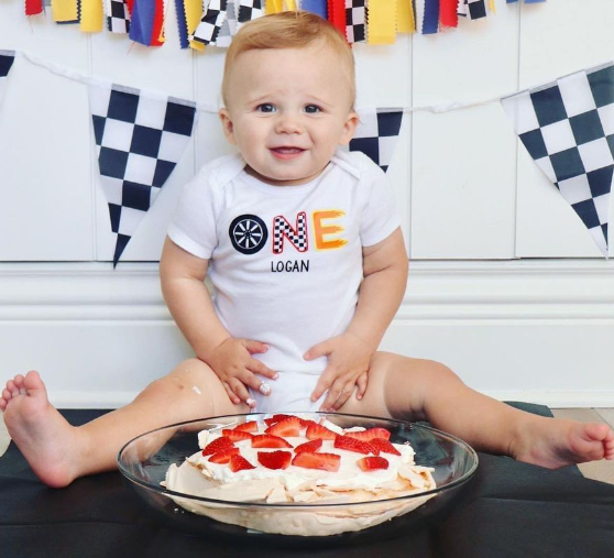 A baby wearing a one shirt is sitting next to a bowl of cake