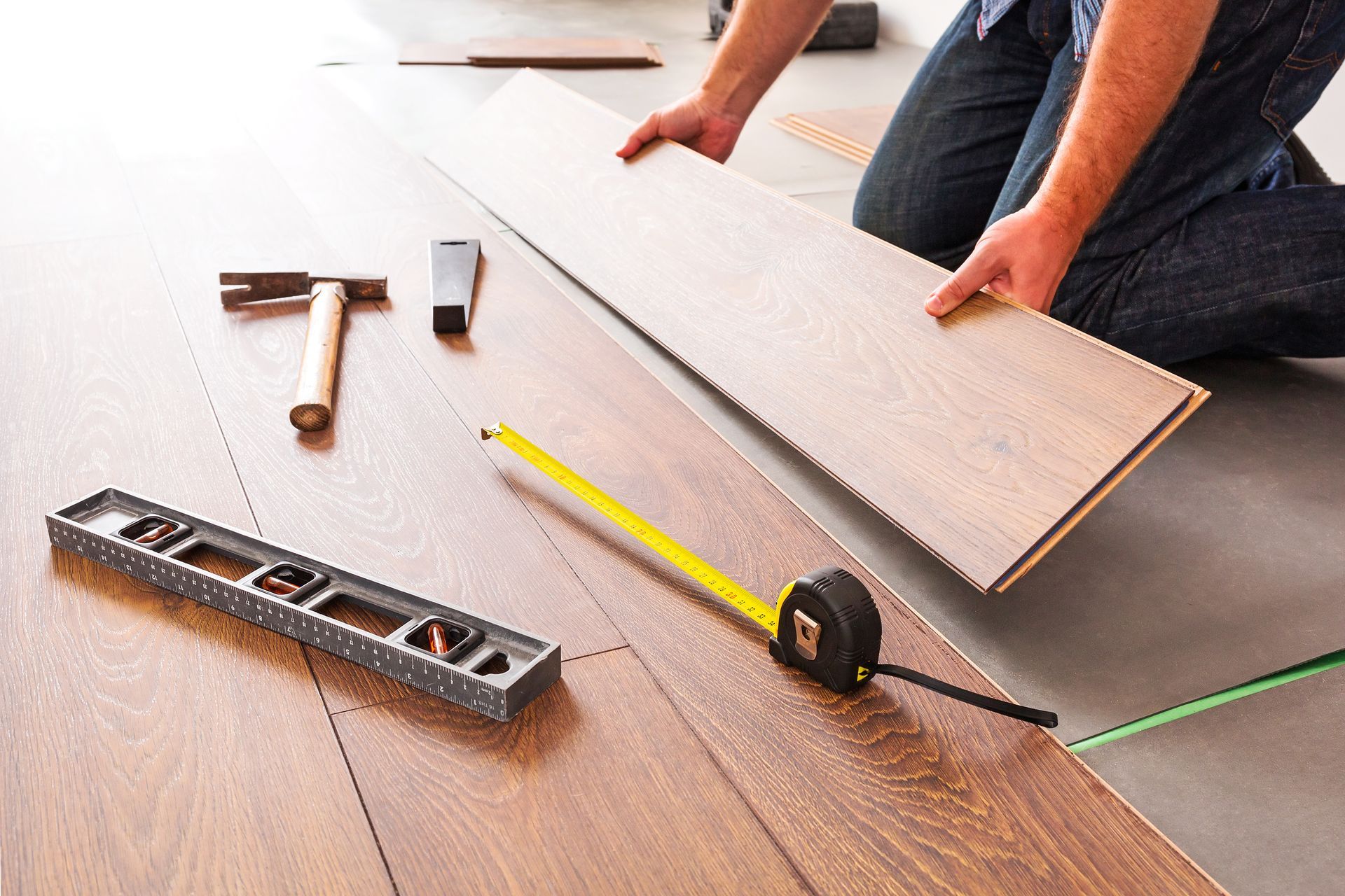 Person laying wood flooring planks on a floor, with a level, hammer, tape measure, and tools nearby.