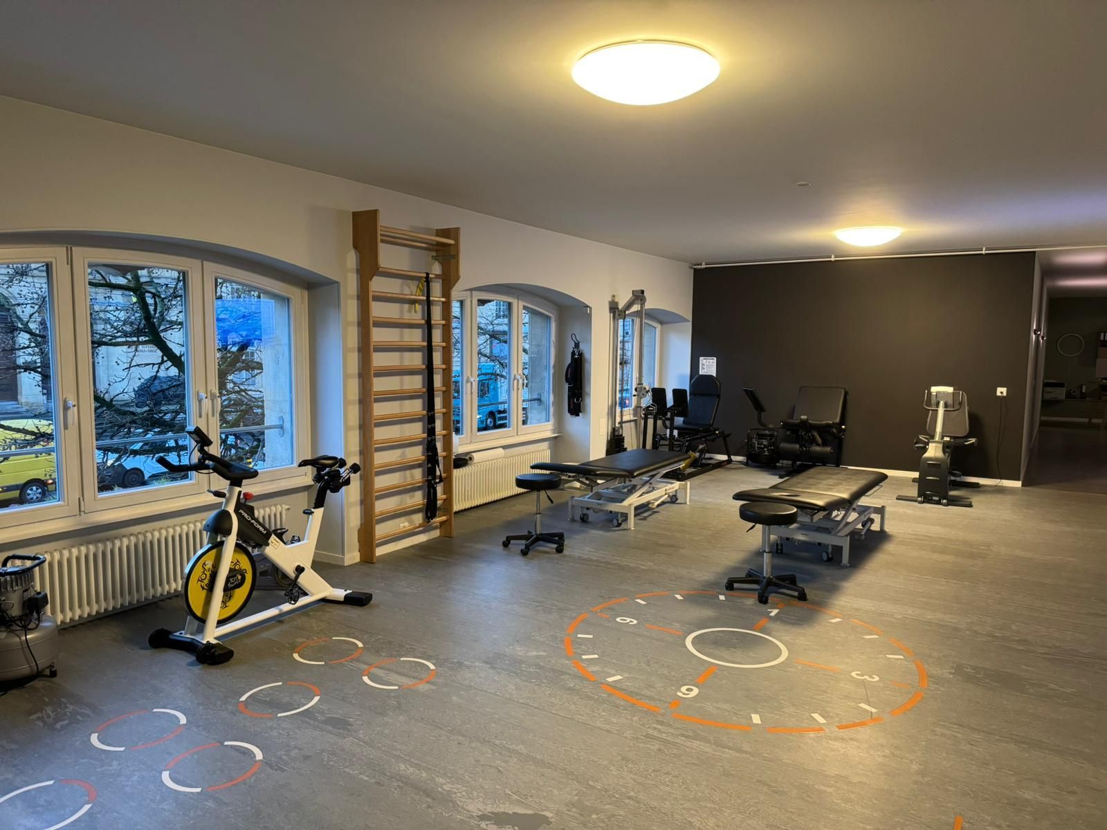Une salle de physiothérapie avec des appareils d'exercice : un vélo stationnaire, des tables de traitement et des barres murales près des fenêtres.