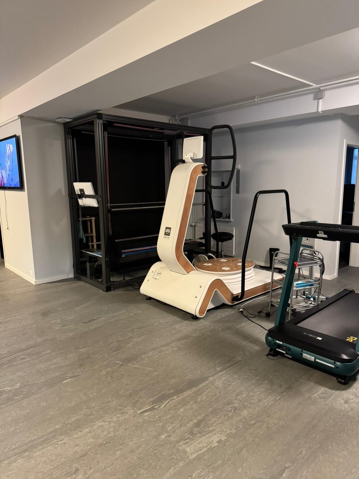 Une salle de sport avec des équipements : une machine blanche et marron, un tapis de course et une structure noire. Le sol est gris et les murs blancs.