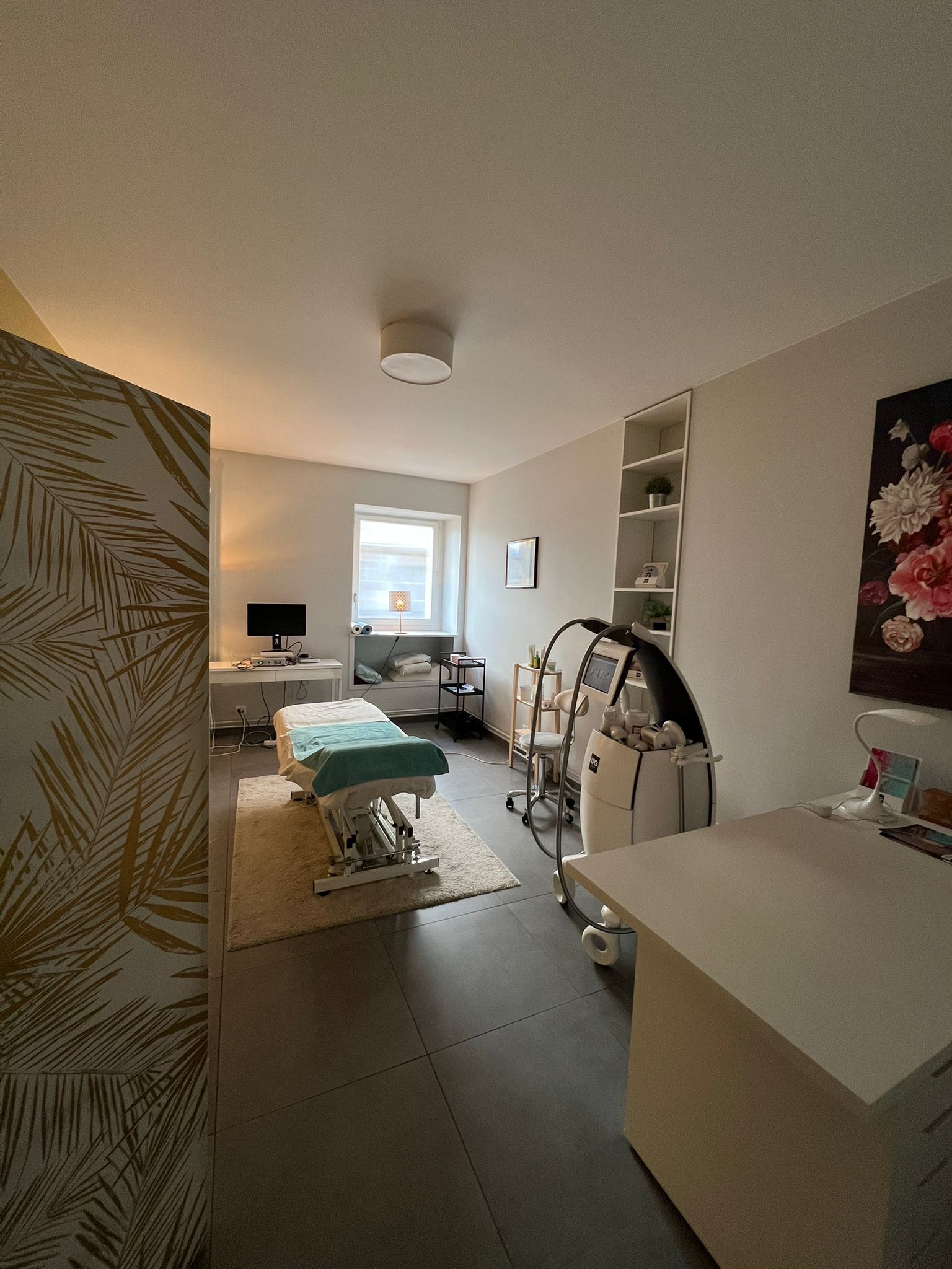 Une salle de soins spa avec table de massage, matériel médical et étagères. La pièce est dotée d'une fenêtre, d'une décoration florale et d'un parquet clair.