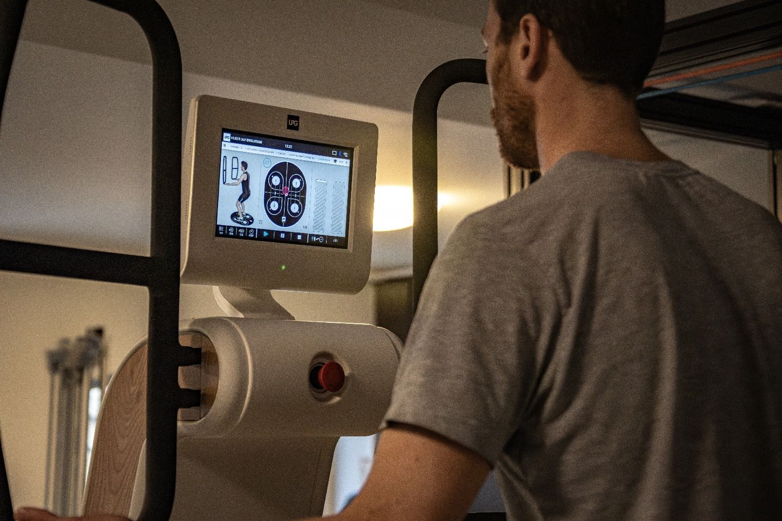 Un homme utilise un appareil d'exercice blanc avec écran, regardant l'écran. Il se trouve dans une pièce avec fenêtre et autres équipements.