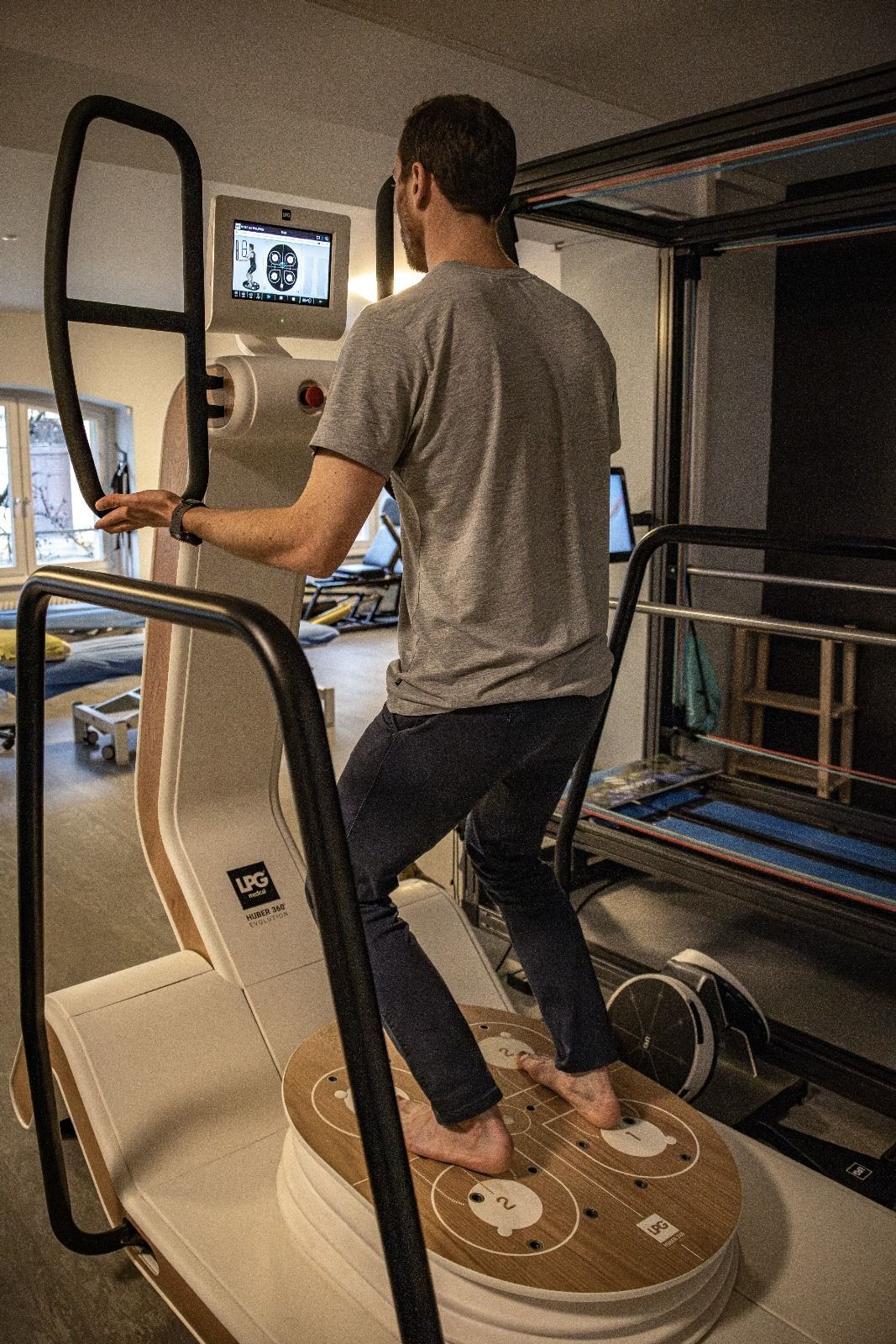 Un homme se tient en équilibre sur une plateforme en bois, en s'appuyant sur des rampes, à l'intérieur. Il porte un jean et une chemise grise.