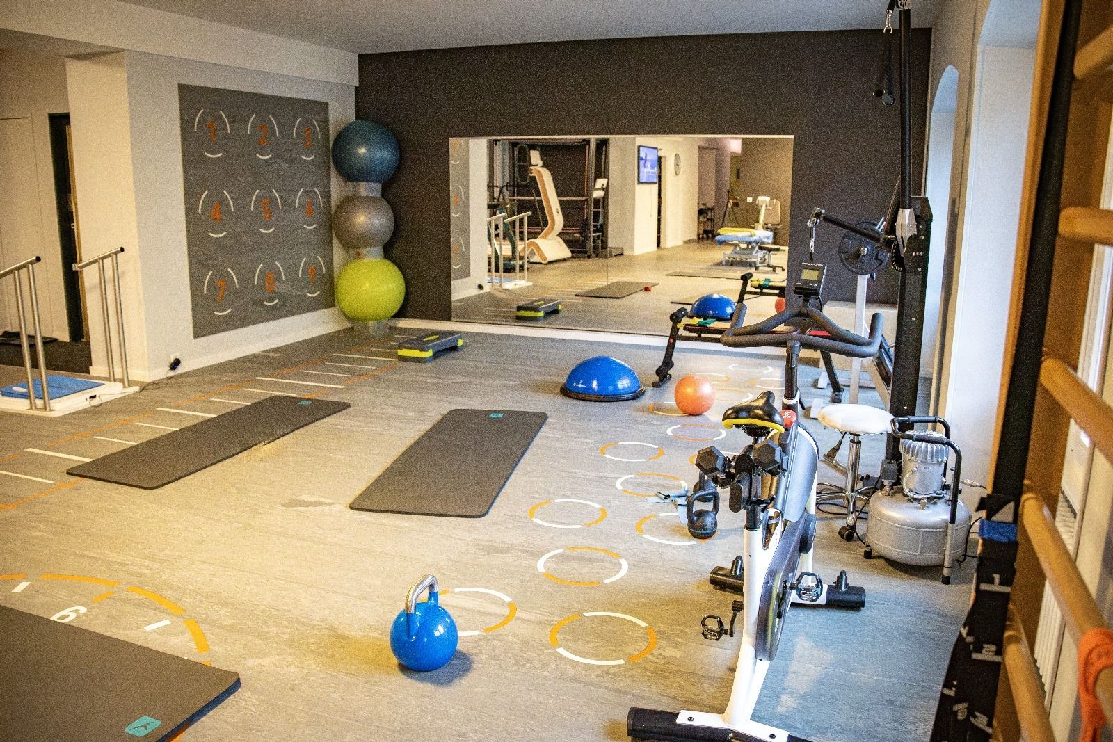 Salle de sport avec appareils d'exercice, tapis et mur miroir. Ballons de yoga et haltères sont disséminés au sol.