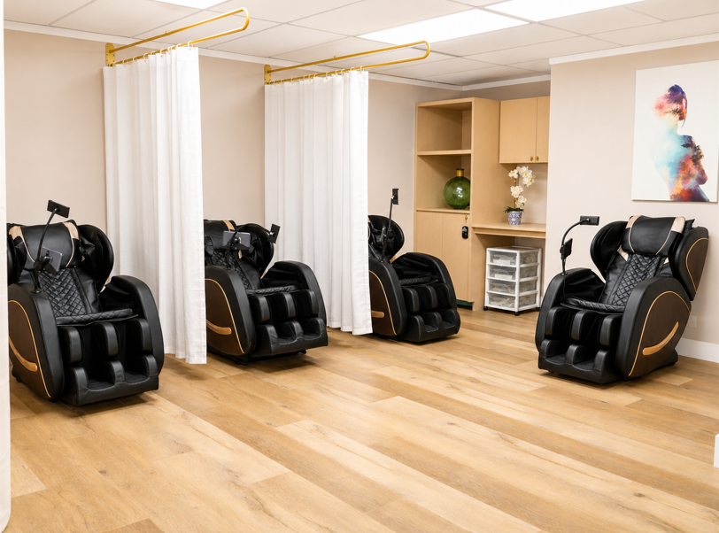 Modern spa room with four black massage chairs, light wood floors, and white curtains