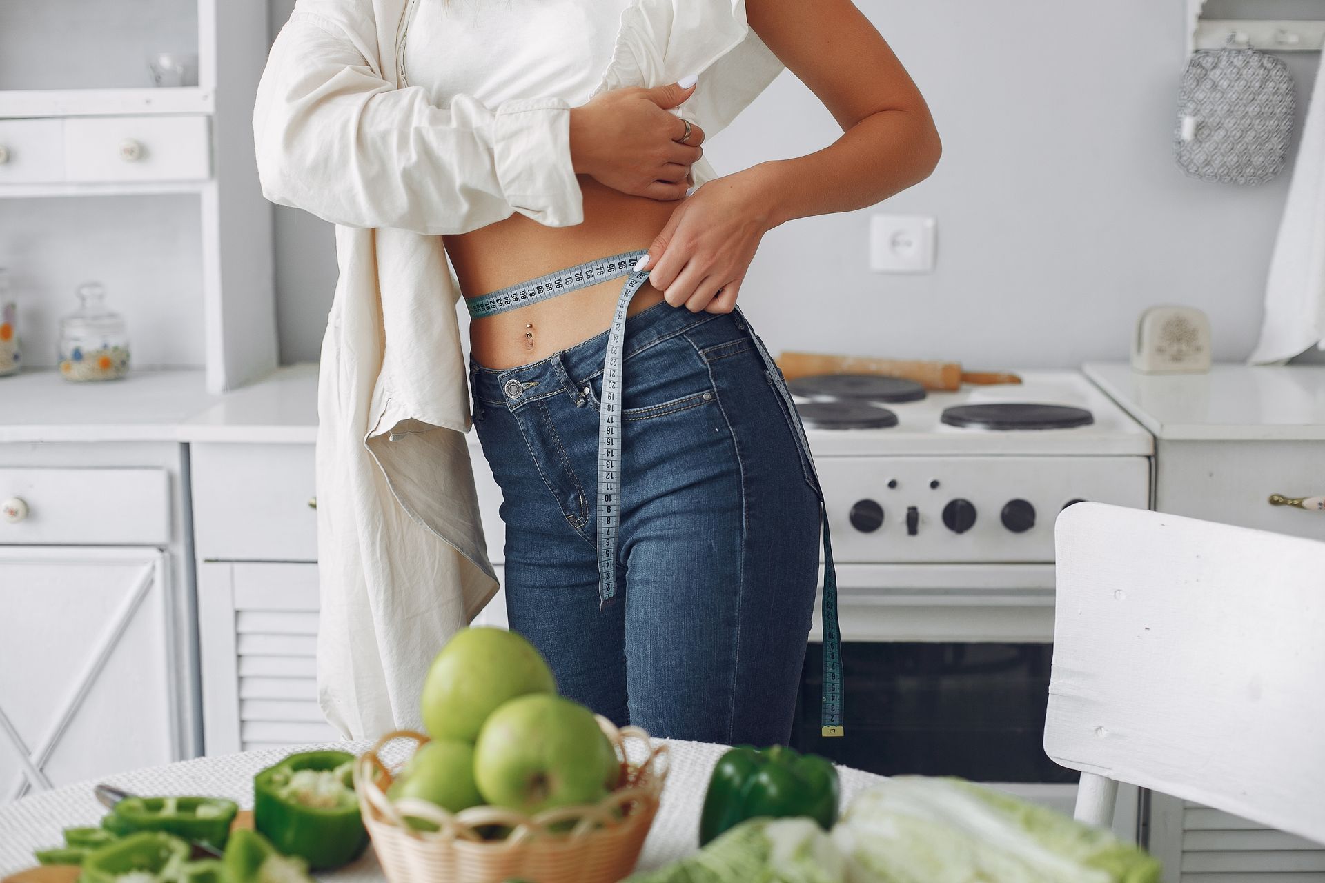 Person buttoning jeans in a bright kitchen, with fresh vegetables on the counter.
