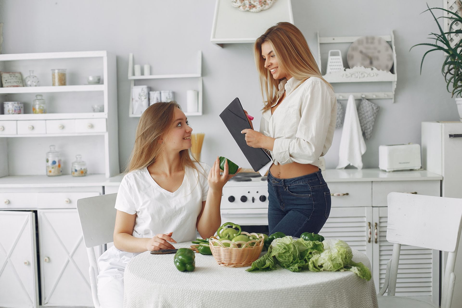 Two women in a bright kitchen sharing ingredients at a table with fresh vegetables.