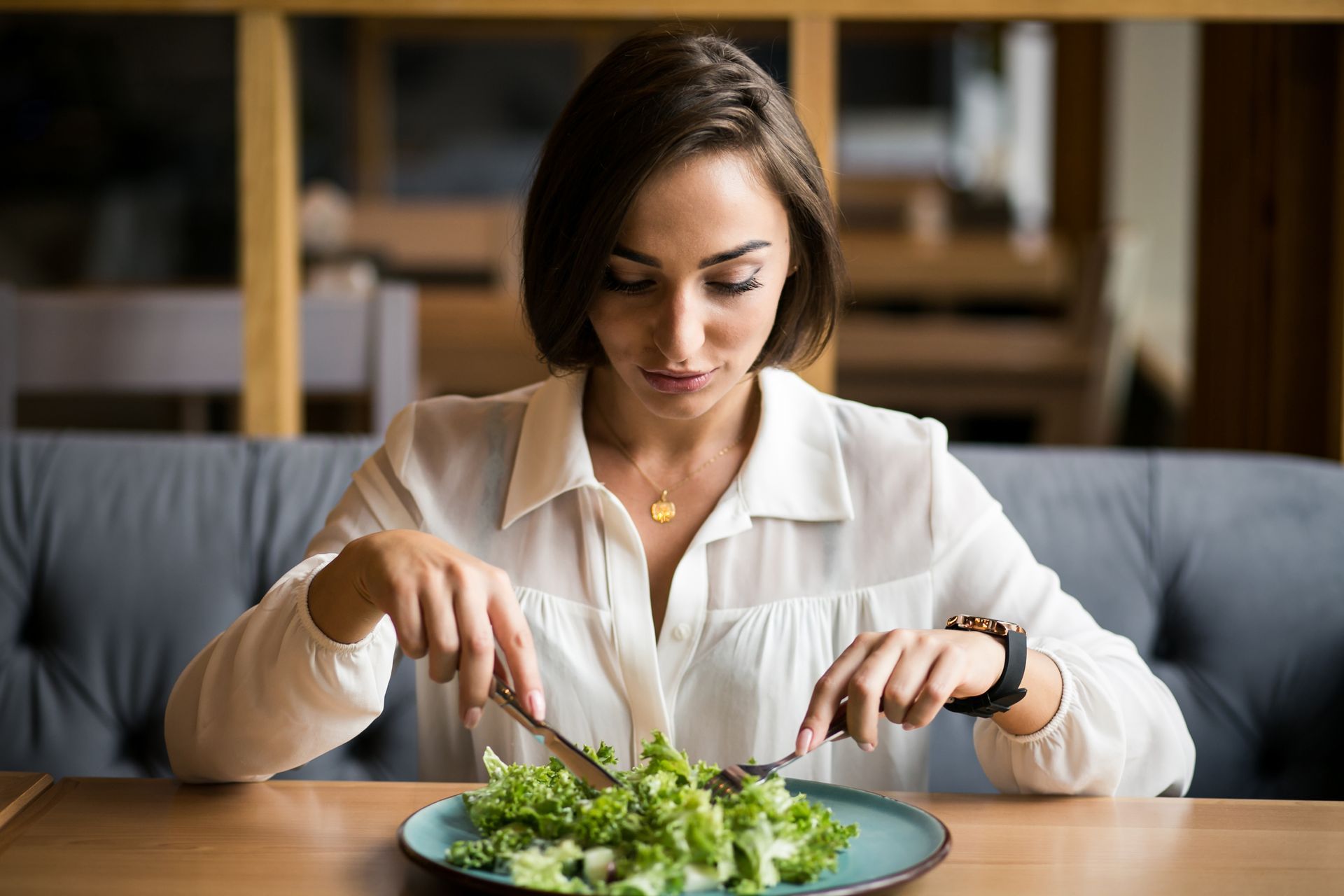Woman eating salad at a table in a cozy cafe, with a fork and knife.