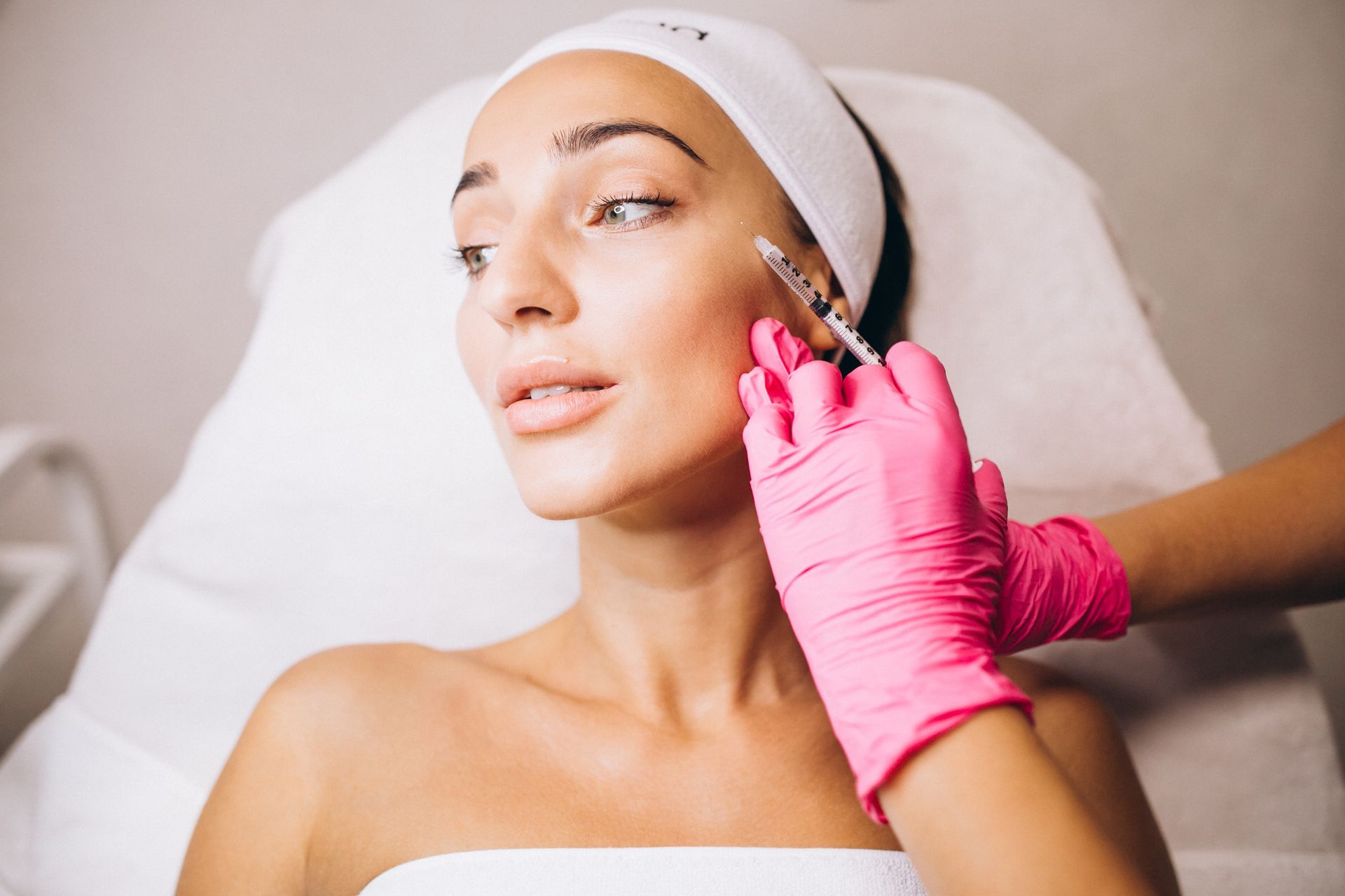 Facial treatment with pink-gloved hands applying serum to a woman’s cheek while she reclines in a spa chair