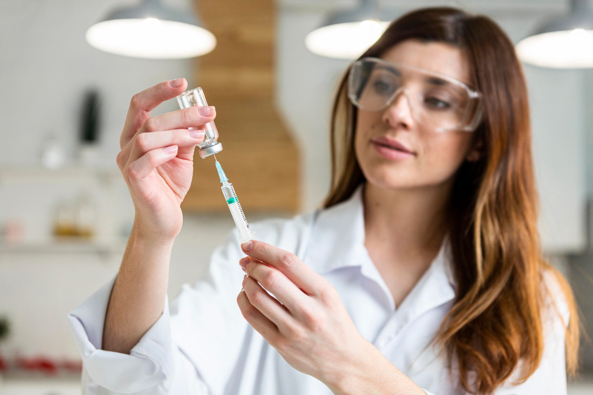 Woman in lab goggles holding a syringe and vial in a bright laboratory.
