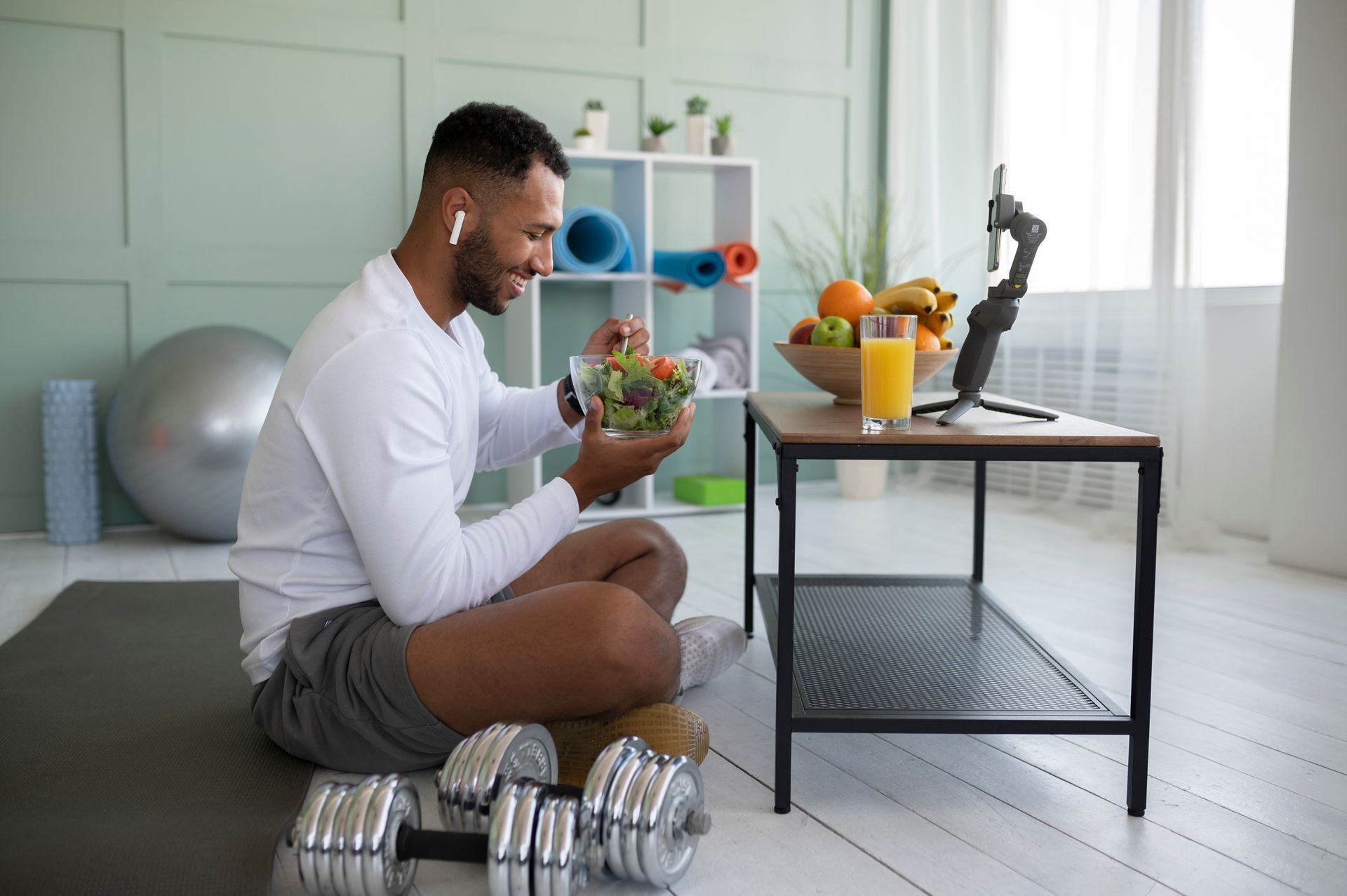 Person sitting on the floor with dumbbells and a salad bowl beside a workout bench in a bright home gym