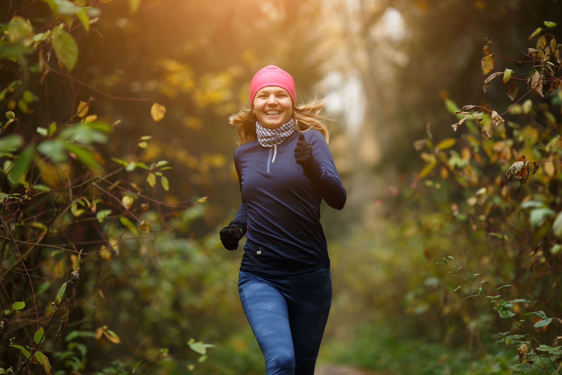 Person jogging on a forest trail, wearing a pink hat and navy top, with sunlight filtering through trees