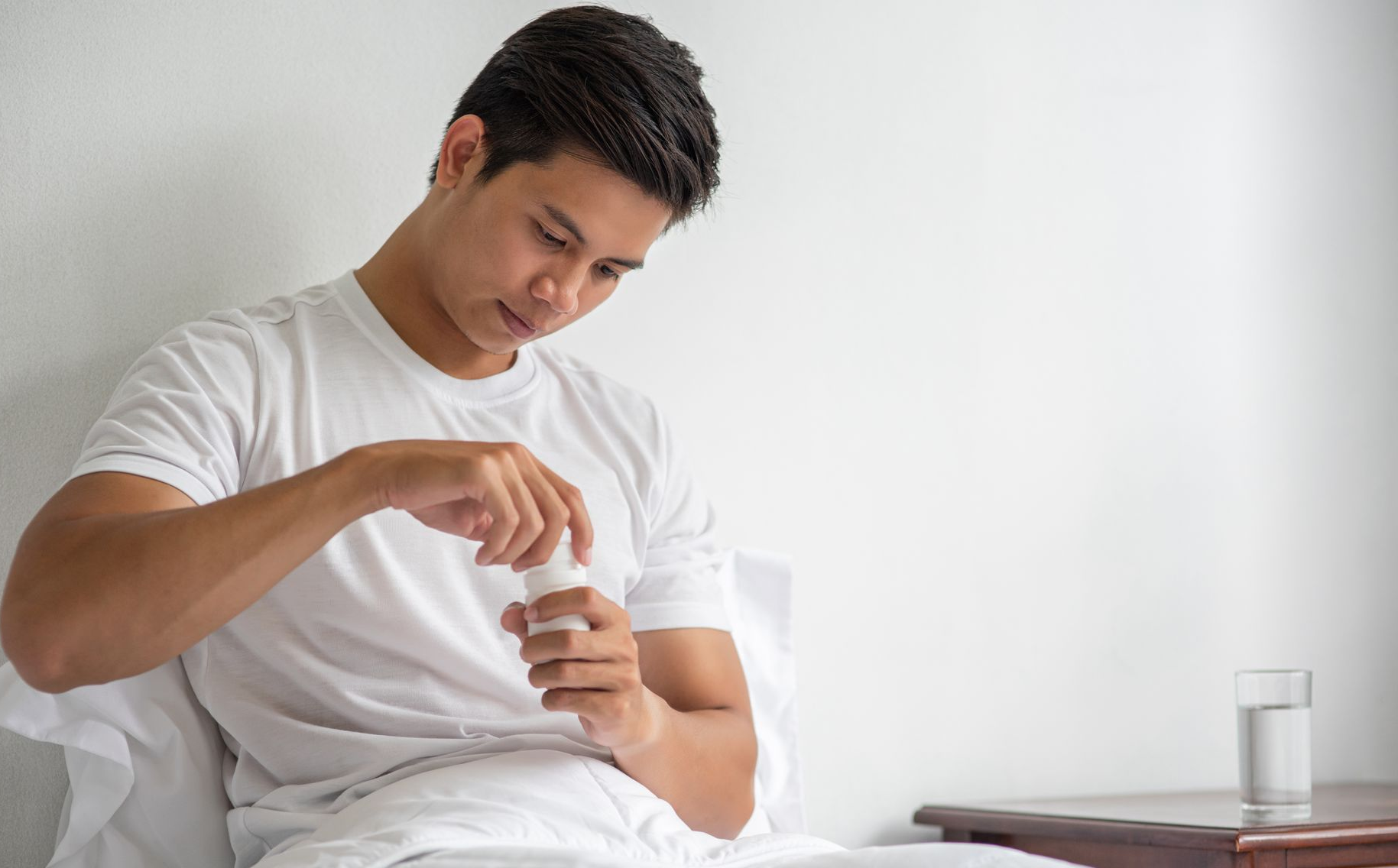 Man in a white shirt opening a medication bottle while sitting on a bed beside a glass of water
