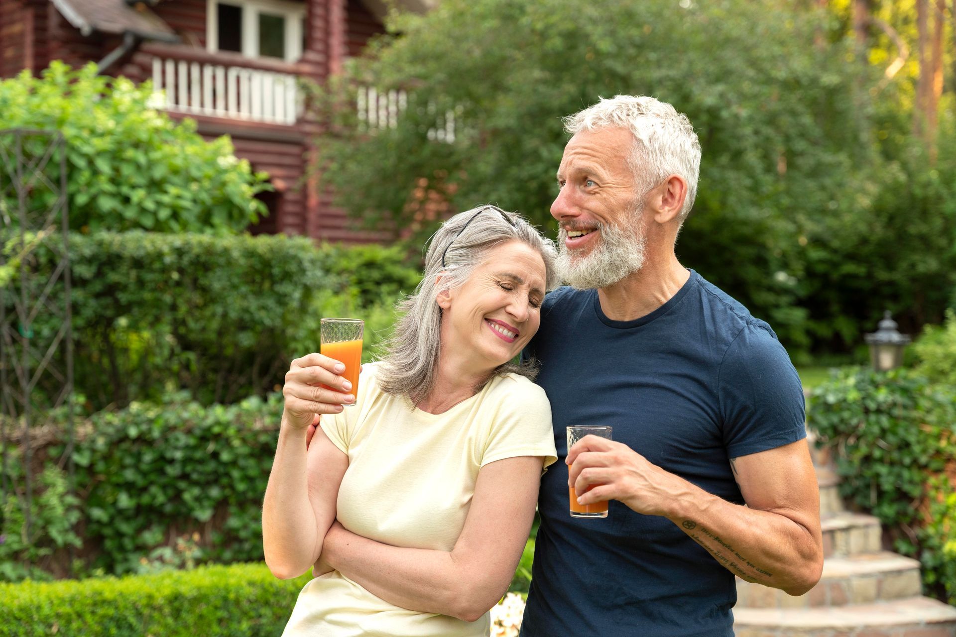 Two smiling adults standing outdoors in a garden, holding drinks and leaning together.