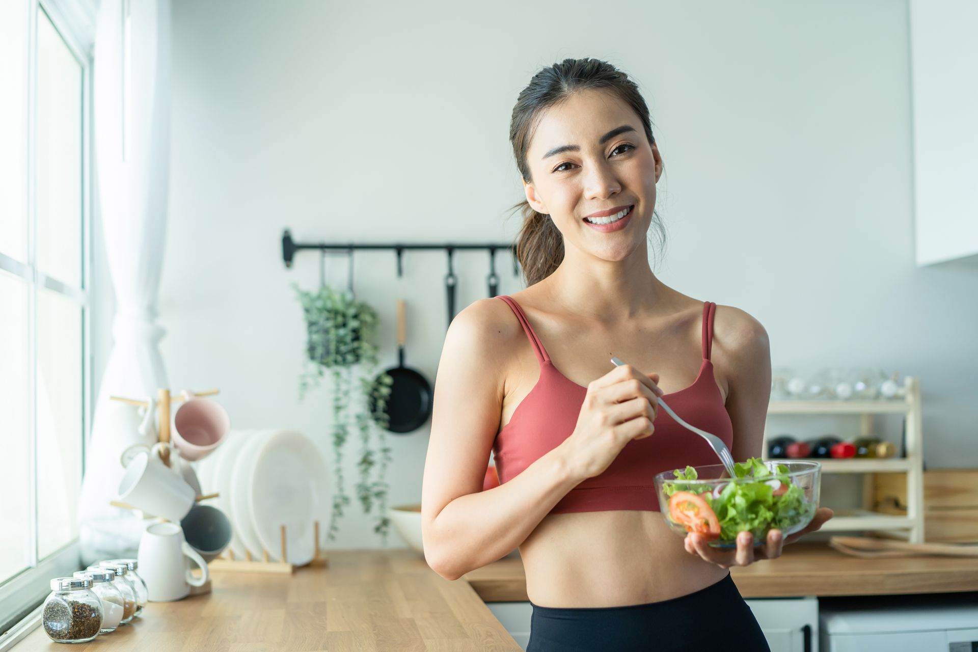 Smiling person in a kitchen holding a bowl of salad, wearing a red sports bra.