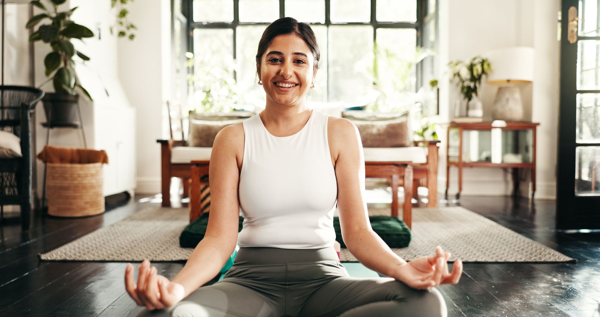 Woman meditating cross-legged on a yoga mat in a bright living room, smiling gently.