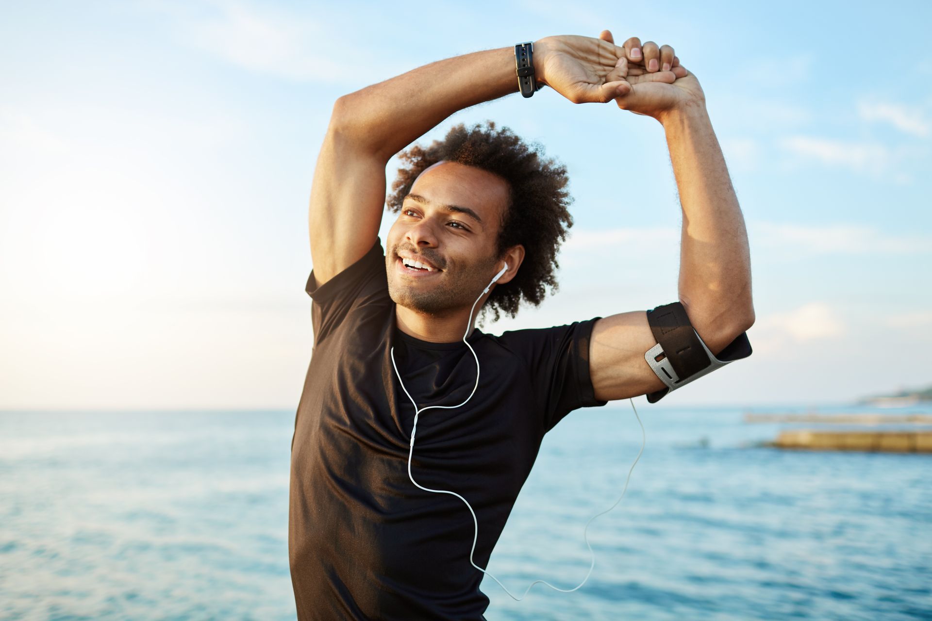 Person stretching on a beach, smiling with arms raised near the ocean at sunset.