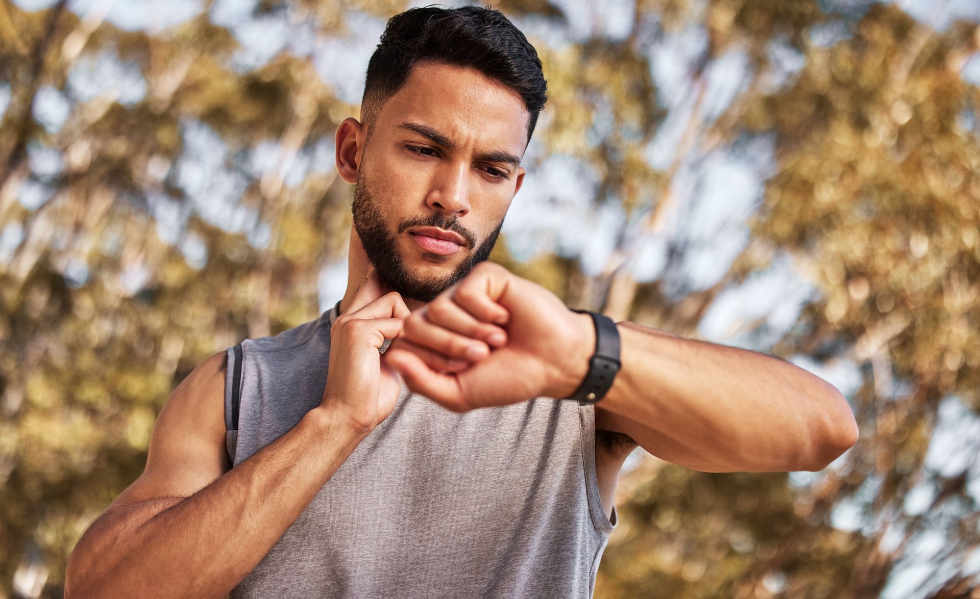 Man outdoors checking his wristwatch, wearing a gray sleeveless shirt against a blurred background
