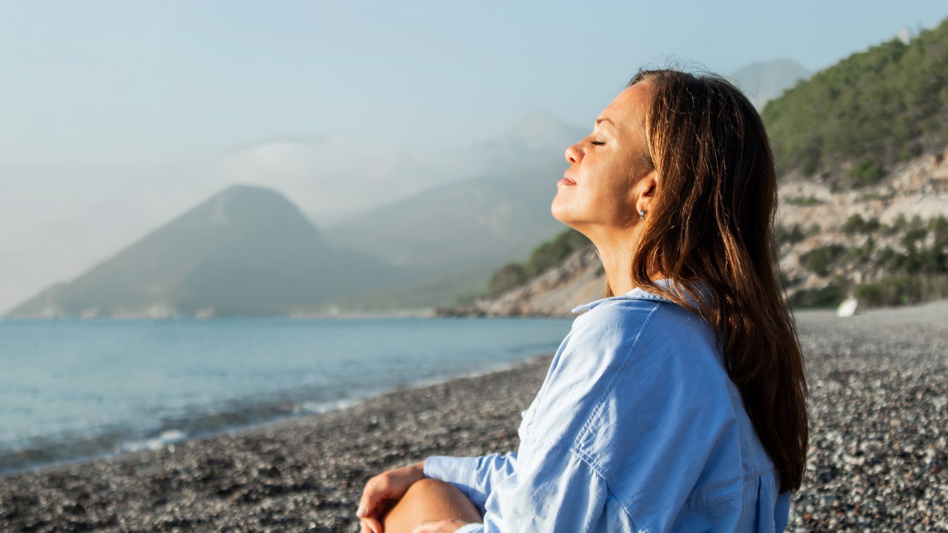 Person sitting on a rocky beach, smiling toward the sun with blue water and mountains in the background